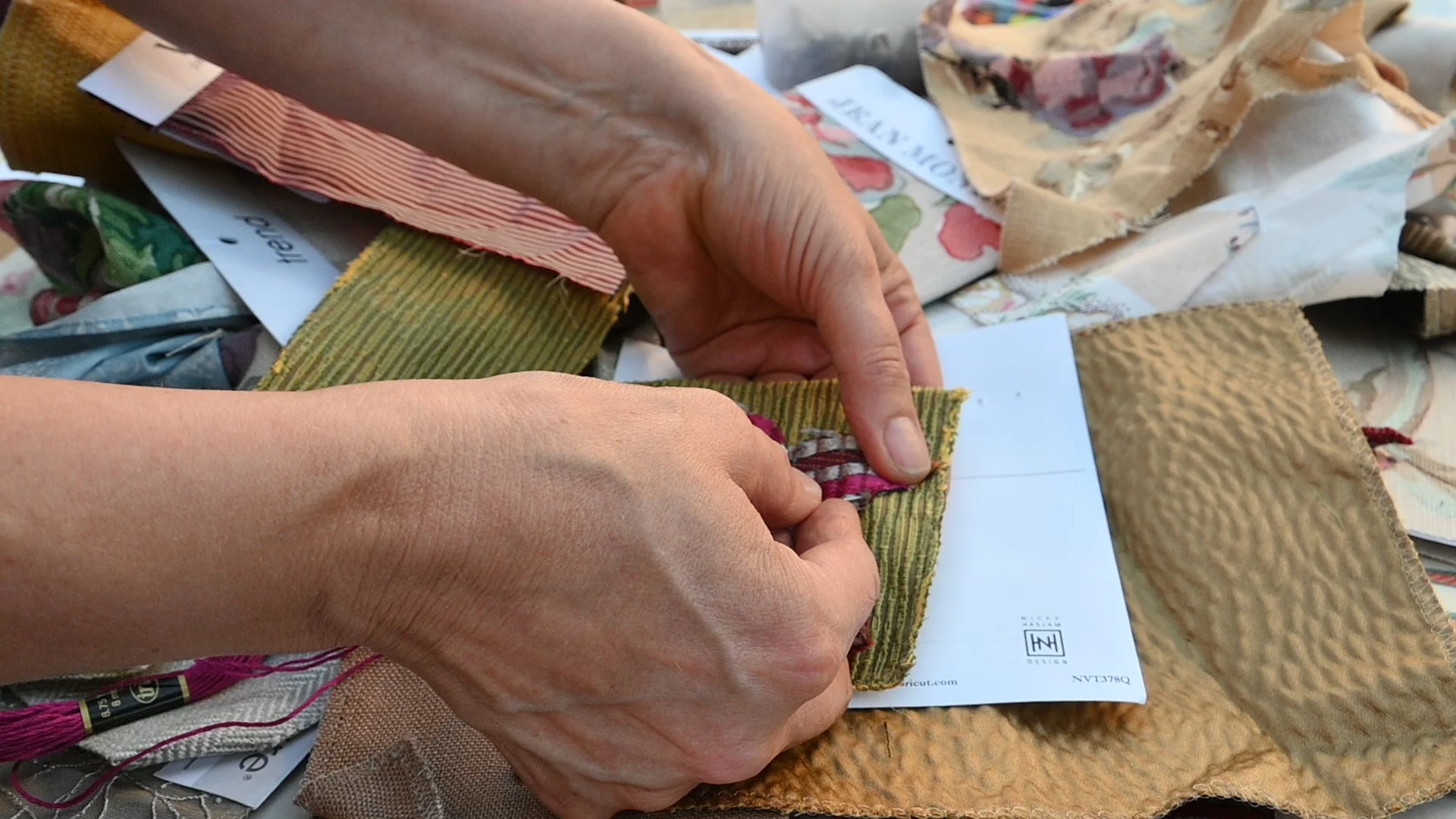 Hands working with fabric swatches and design samples, possibly selecting or arranging fabrics for a craft or project.
