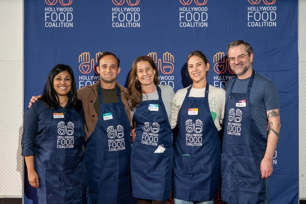 Group of five people standing in front of a blue backdrop with the Hollywood Food Coalition logo. They are wearing aprons with the same logo and appear to be volunteers at a food charity event.