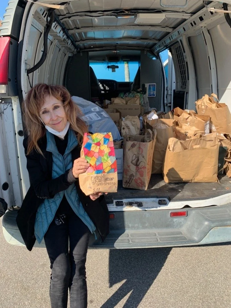 A woman with red hair wearing a face mask around her neck, standing next to an open van filled with paper grocery bags. She holds a paper bag with colorful geometric shapes and the words 'Sunflower Seed Spread' written on it.