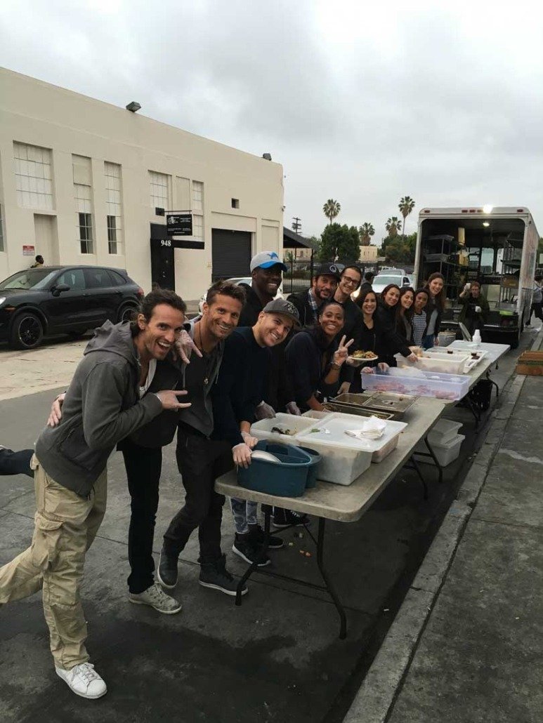 A group of people standing behind a serving table outdoors, with food containers and a food truck, under cloudy skies.
