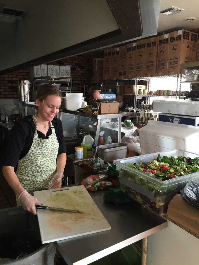 A smiling woman wearing a floral apron and gloves preparing food at a kitchen counter with a large container of salad and various kitchen supplies around her.