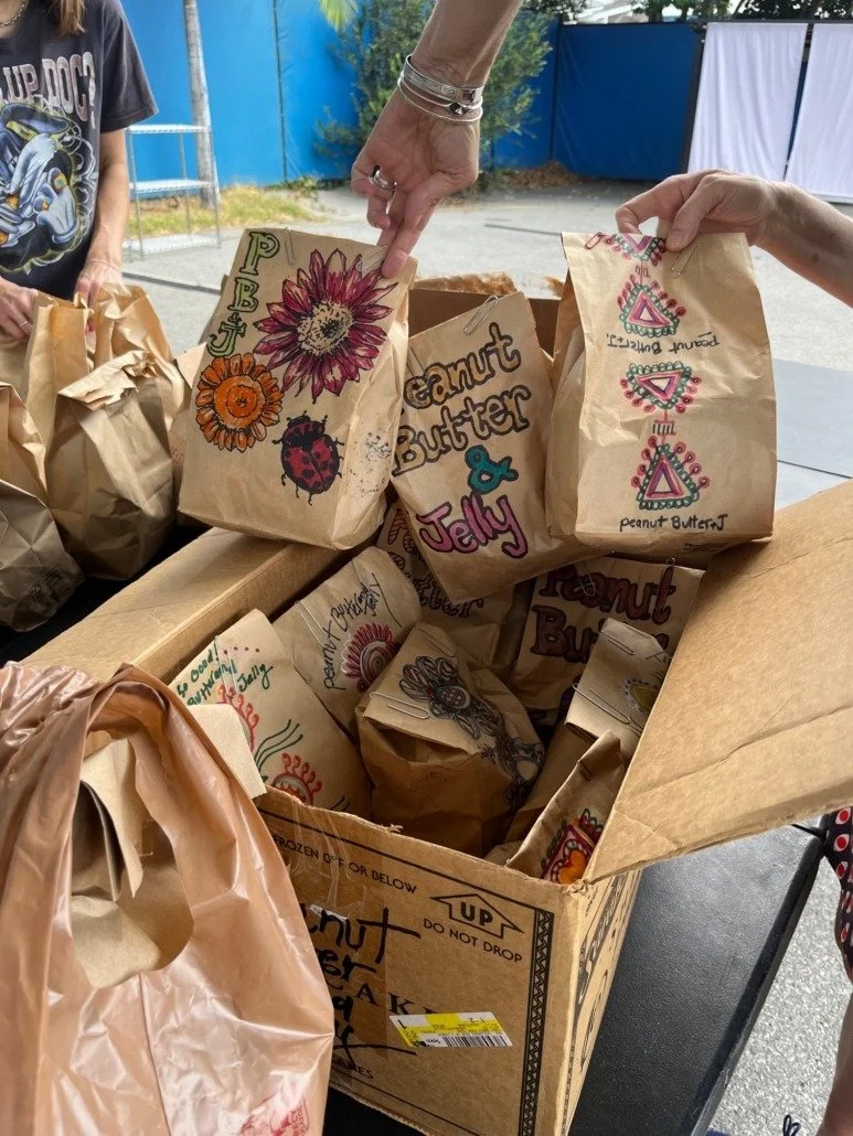 Colorful paper bags with hand-drawn designs and labels, placed in a large cardboard box, with two people reaching in to pick them up.