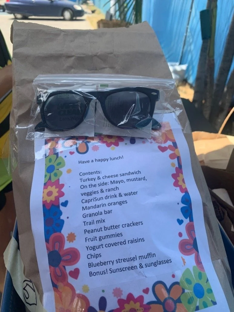 A paper bag with a colorful floral border containing a lunch menu and a pair of sunglasses in a plastic bag on top.