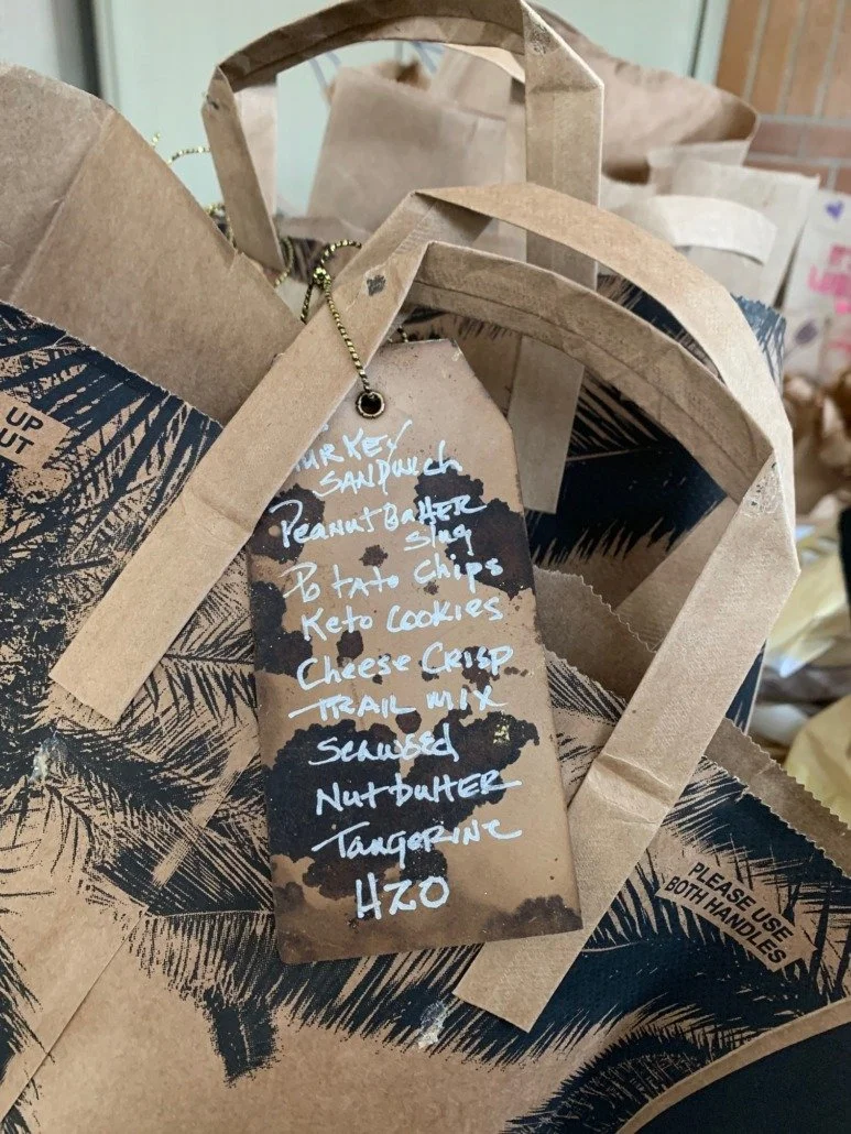 Paper gift bags with a list of food items written on a hanging tag, including turkey sandwich, peanut butter sling, to tater chips, keto cookies, cheese crisp, trail mix, scrambled nut butter, tangerine, and HZO.