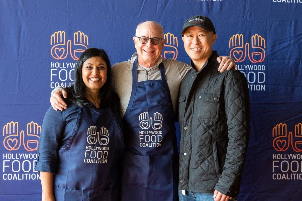 Three people standing in front of a blue backdrop with the Hollywood Food Coalition logo, smiling and posing for the photo, with two wearing aprons and one in a jacket and hat.