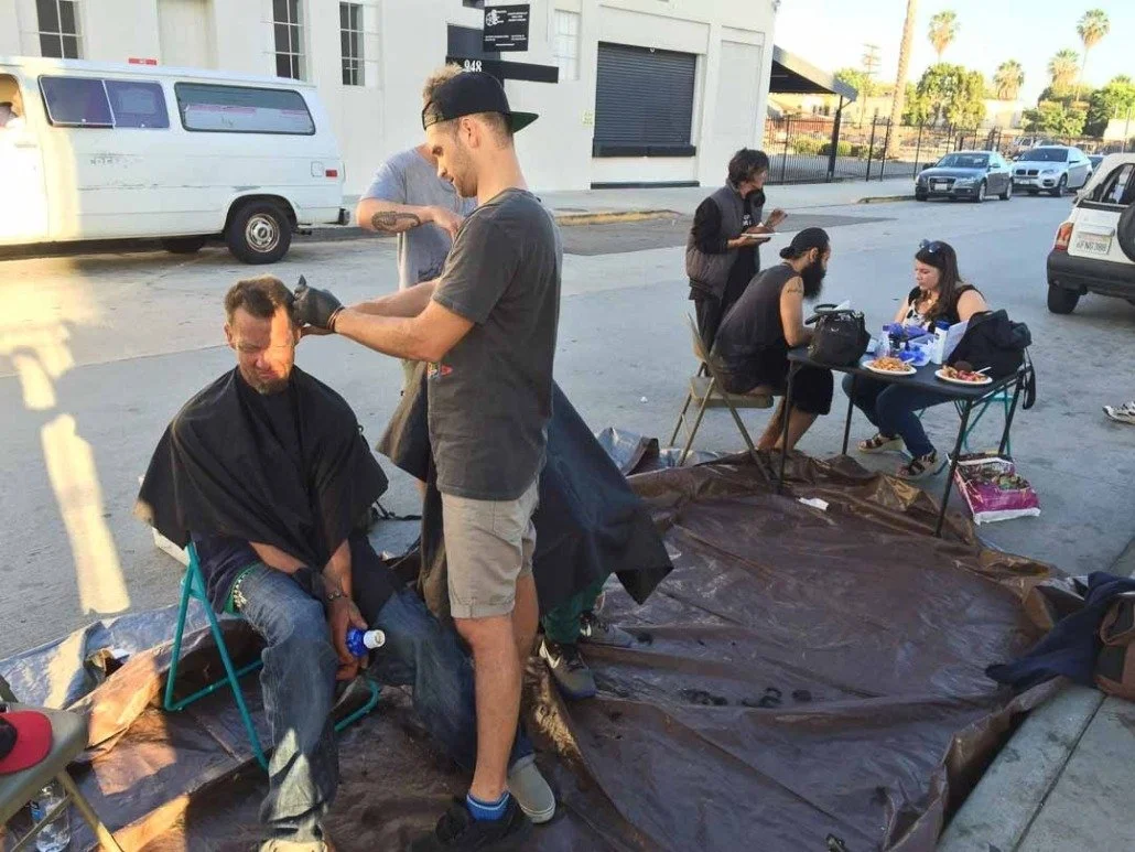 A man receives a haircut outdoors on the street, with a tarp on the ground, while others sit at a table eating nearby.