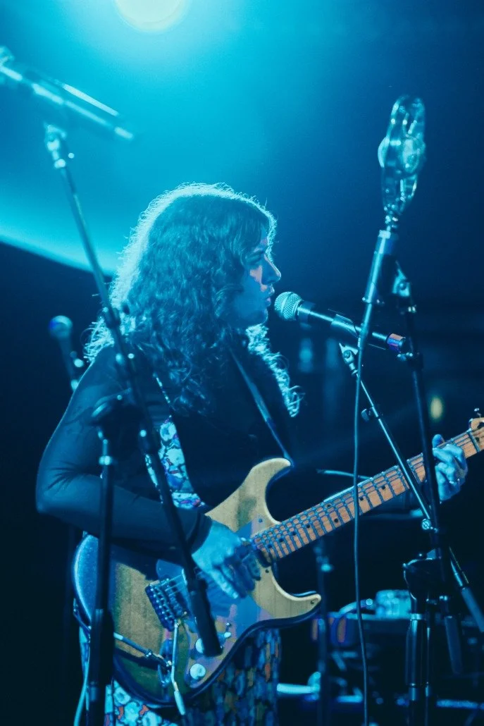 A woman with curly hair playing an electric guitar and singing into a microphone on stage with blue lighting.