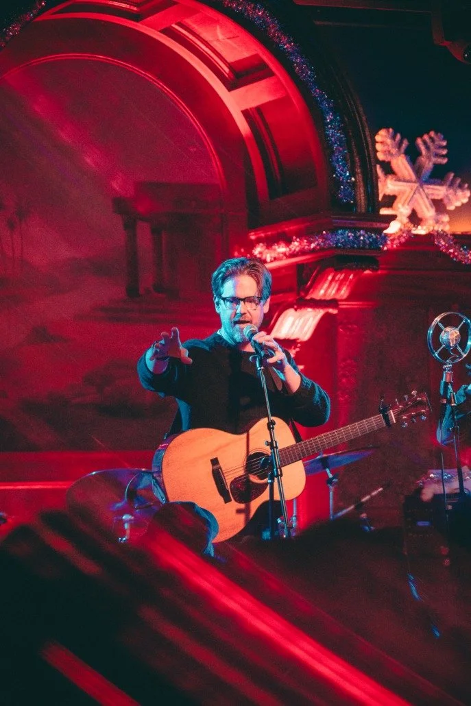 A man with glasses holding a guitar and singing into a microphone on stage, decorated with holiday lights and a large snowflake decoration in the background.