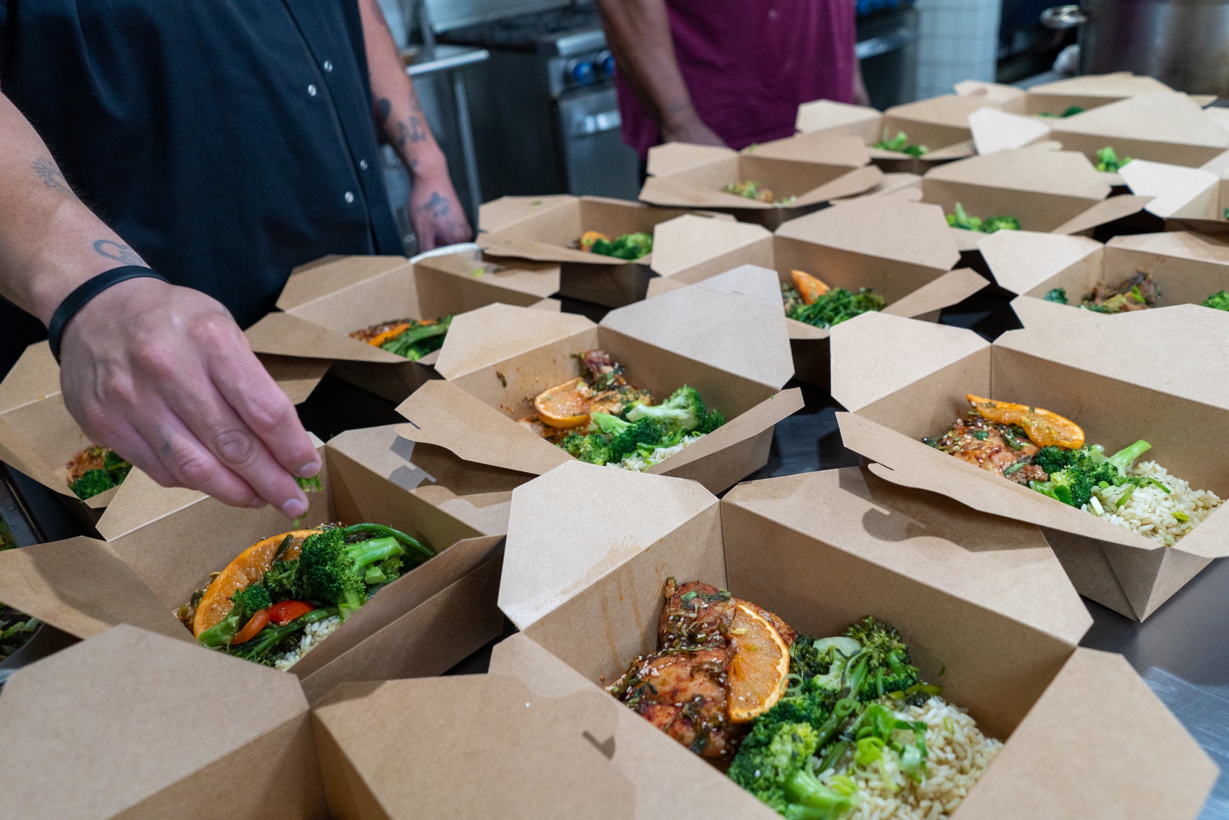 Multiple brown takeout boxes filled with rice, vegetables, and grilled chicken, on a stainless steel table in a restaurant kitchen, with people preparing and packing food.