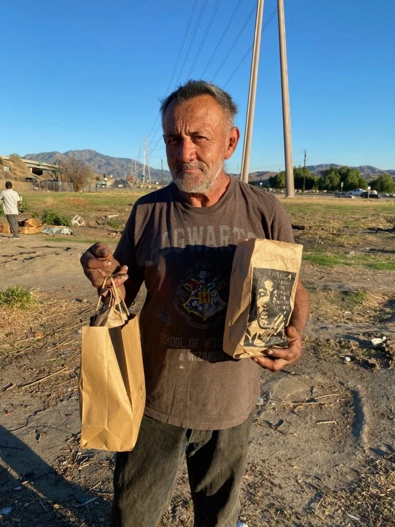 An elderly man with a gray beard and short hair standing outdoors during daylight, holding paper bags, with mountains and power lines in the background.