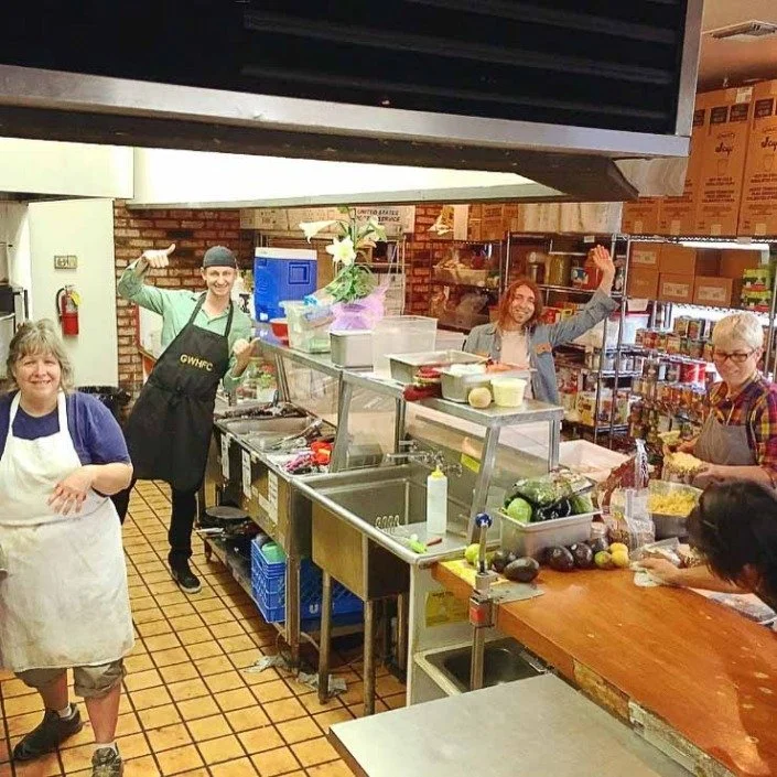 Four women working in a busy kitchen or food preparation area, some smiling and waving at the camera.