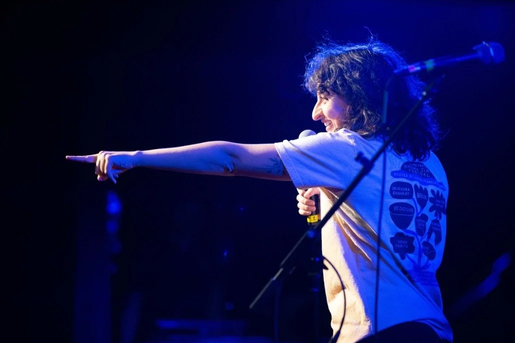 A woman with curly hair on stage, smiling, pointing to the left, holding a microphone, illuminated by blue stage lighting.