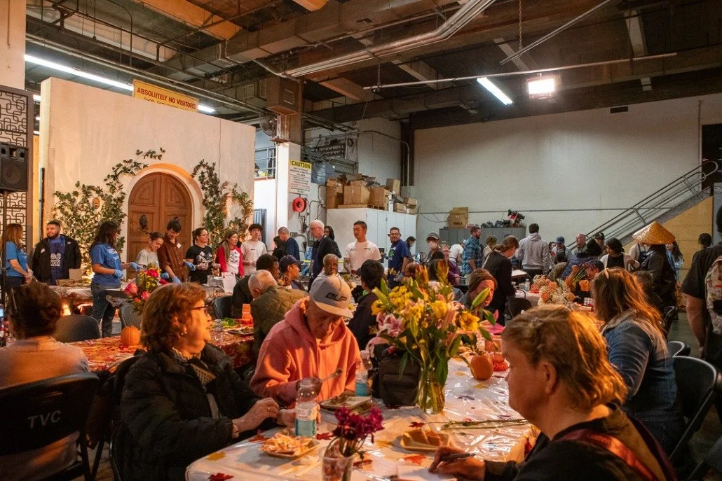 People gathered around tables decorated with pumpkins and flowers in a spacious indoor event space with high ceilings, industrial lighting, and cardboard boxes stacked in the background.