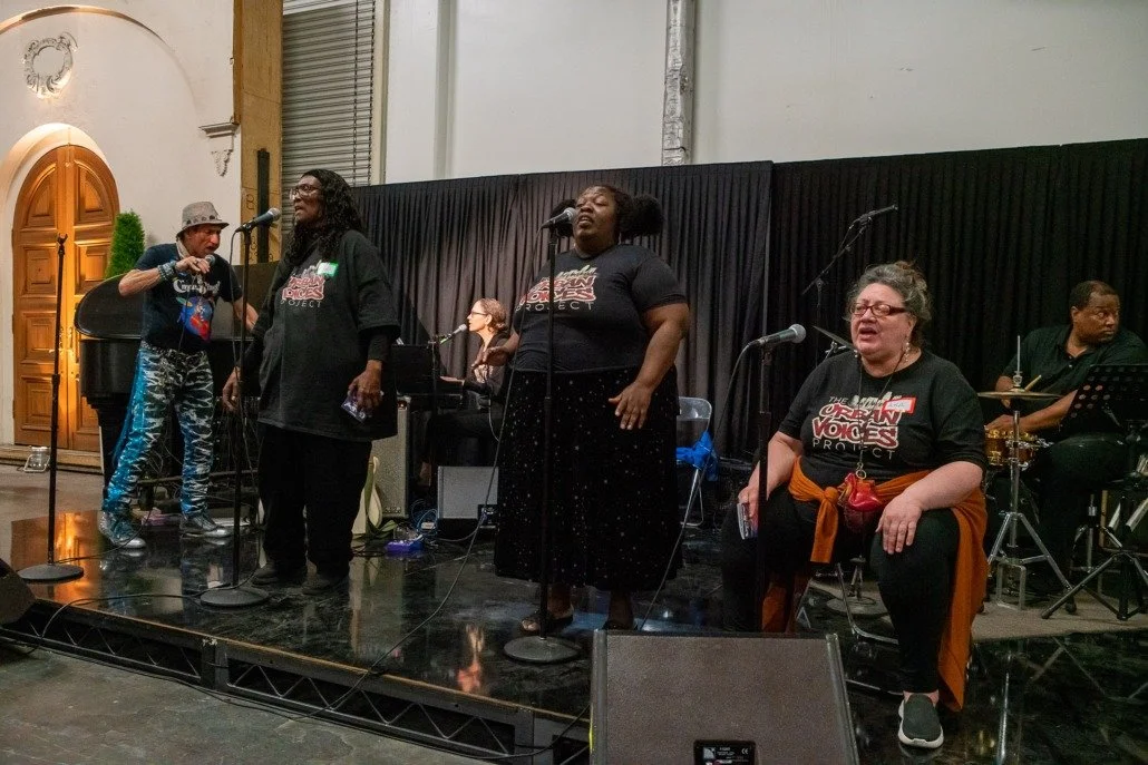 A group of diverse women singing and playing musical instruments on stage at an indoor event or performance.