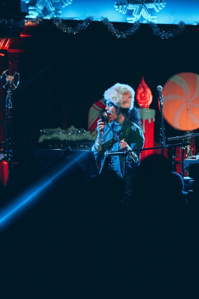 A person singing into a microphone on a stage decorated with Christmas-themed decorations like a large peppermint swirl, a candy cane, ornaments, and holiday garlands, with blue lighting and an audience in the foreground.