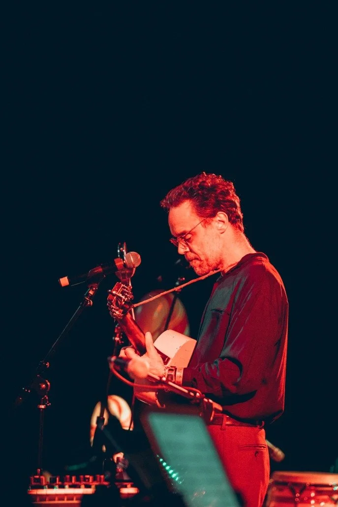 Man with glasses playing an acoustic guitar on stage, with microphone and music equipment nearby, under dim stage lighting.
