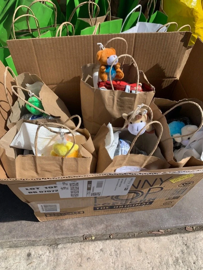 Multiple small paper bags with stuffed animal toys inside, placed inside a large cardboard box on the sidewalk. The stuffed animals include various animals like a bear, a llama, and others. Some bags have small round objects on top, possibly toys or 