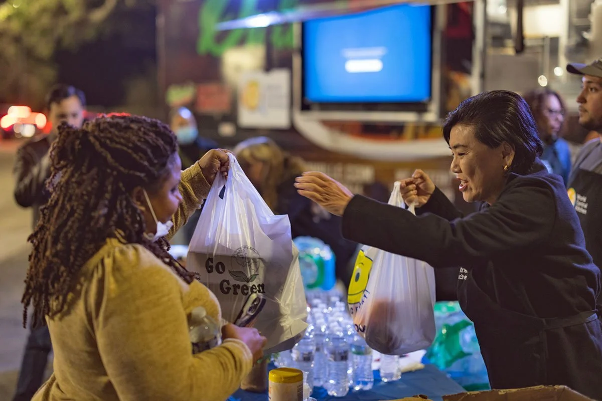 A woman with short dark hair handing a plastic bag to a woman with long dreadlocks and face mask at an outdoor event during evening. There are water bottles on the table and people in the background.