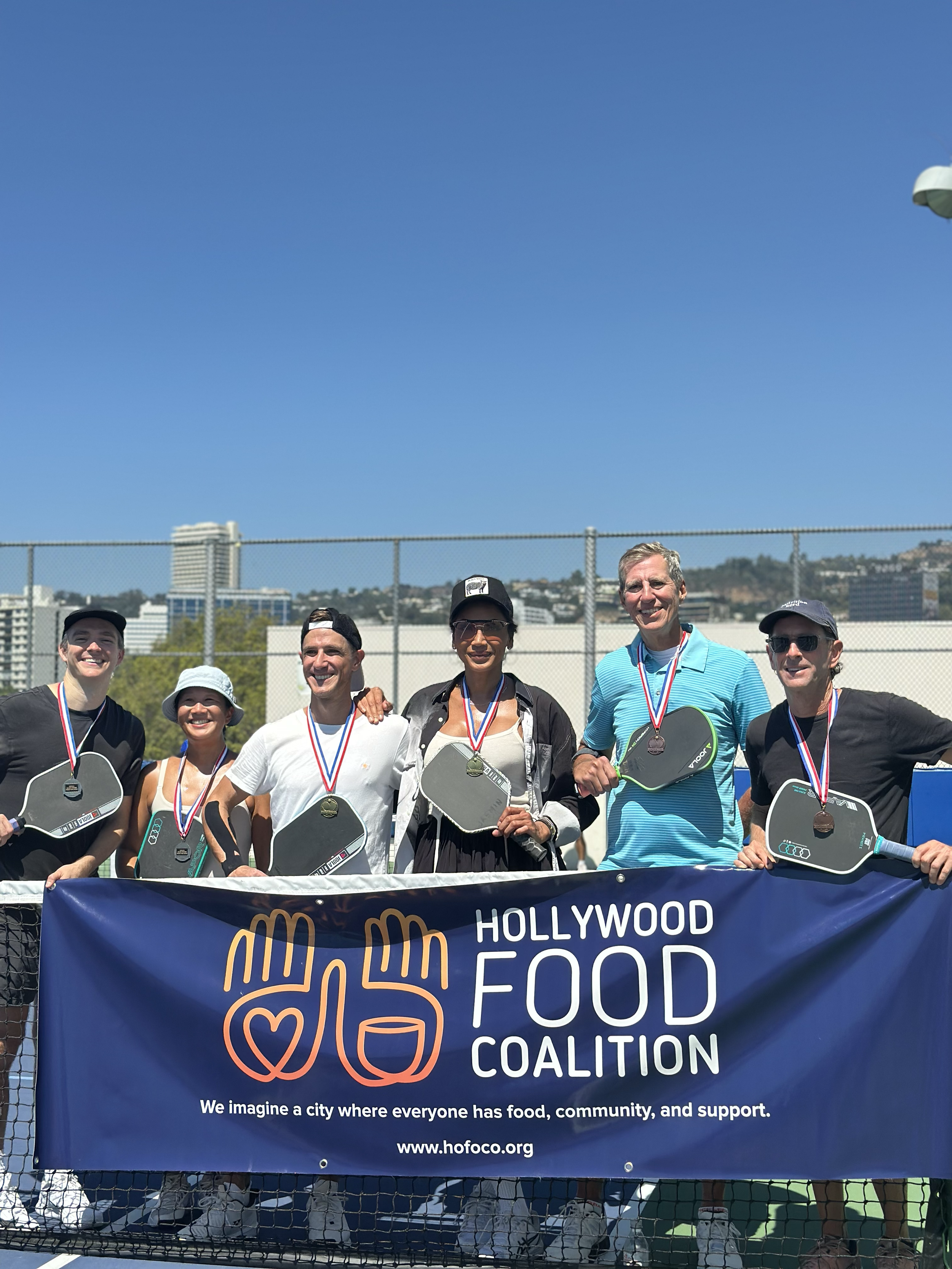 Group of six people on a tennis court holding paddles and wearing medals, standing behind a banner that reads 'Hollywood Food Coalition' with logo and website, during a sunny day.