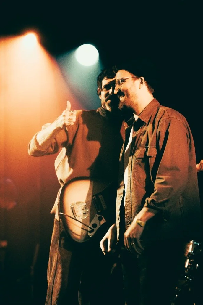 Two men with glasses enjoying a close moment on stage during a concert, one is wearing a brown jacket and the other is wearing a beige jacket, with stage lighting overhead.