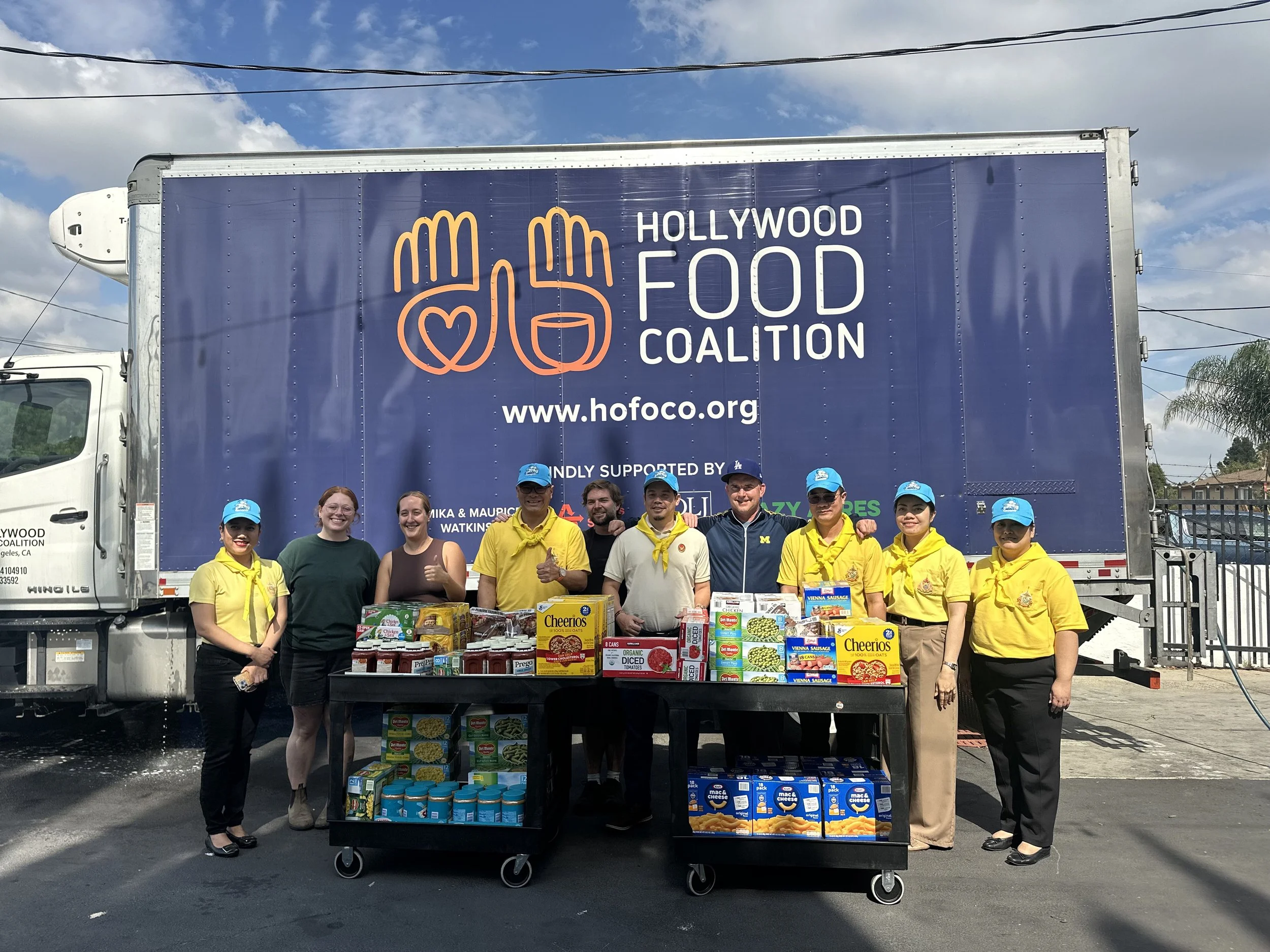 Group of nine people standing in front of a large Hollywood Food Coalition truck, with a cart of canned goods and food items in front of them, outdoors on a sunny day.