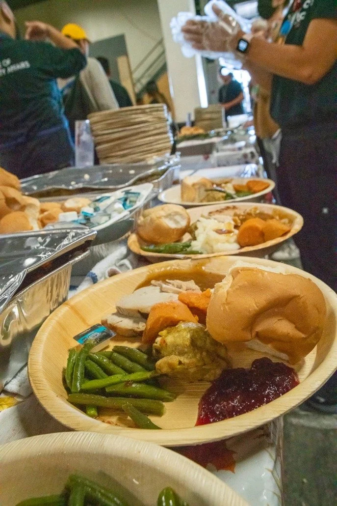 Buffet-style food with green beans, turkey, stuffing, mashed potatoes, bread rolls, cranberry sauce, and gravy on a disposable wooden plate. In the background, there are other plates and people preparing food.