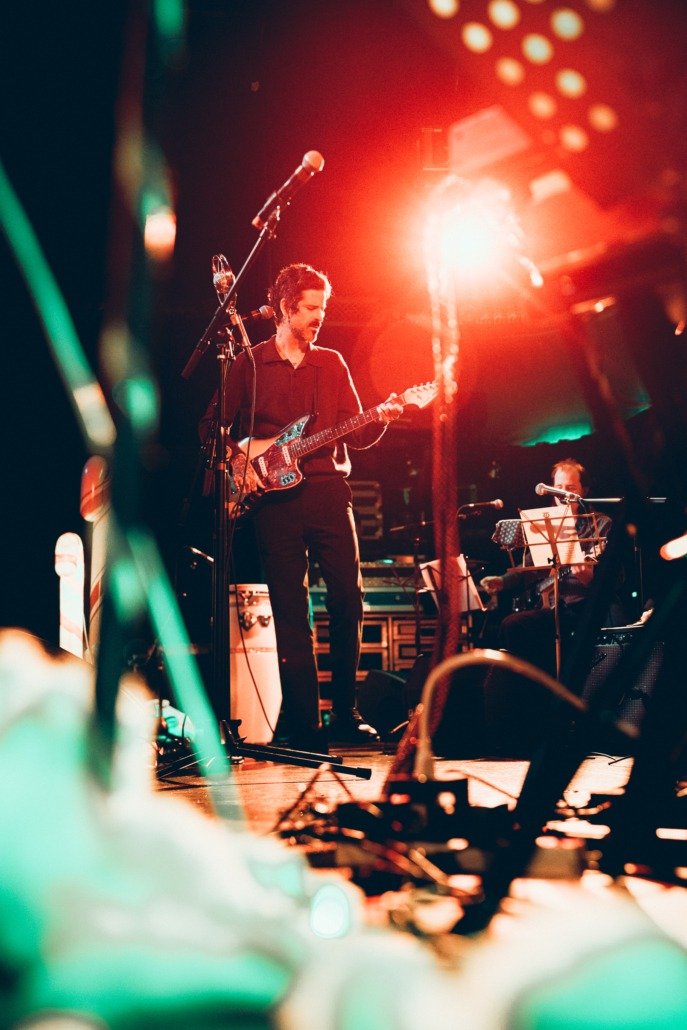 A musician plays an electric guitar on stage under vibrant lighting, with another musician sitting with a keyboard nearby, during a performance or concert.