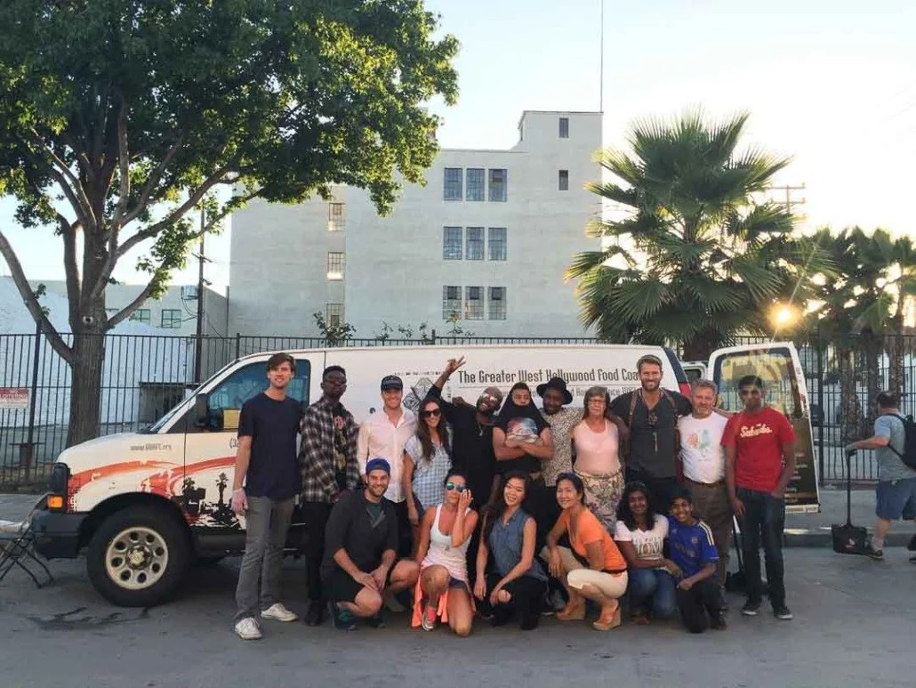 Group of people standing and kneeling in front of a food truck with a tree, palm tree, and building in the background.