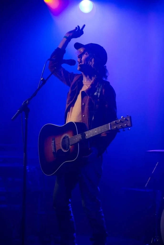 A person performing on stage with a guitar, wearing a baseball cap and a jacket, under colorful stage lighting.