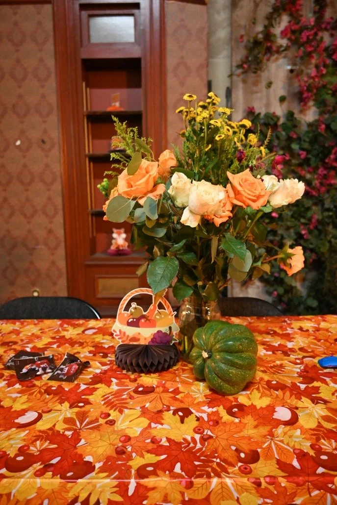 A table decorated with an autumn-themed tablecloth featuring orange and yellow leaves and pumpkins, a large green pumpkin on the right, a bouquet of assorted flowers including roses and greenery in a glass vase, a Halloween-themed paper decoration, a
