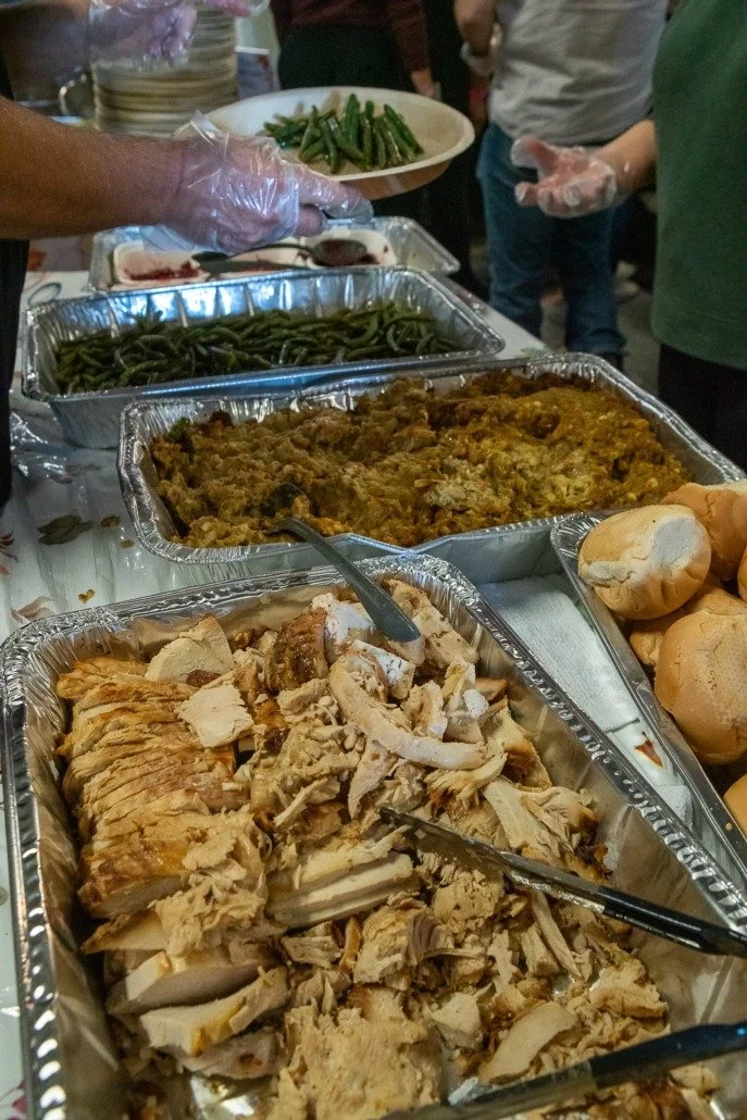 Various dishes of prepared food in aluminum trays, including sliced turkey, green beans, mashed potatoes with gravy, and baked rolls at a buffet-style meal.