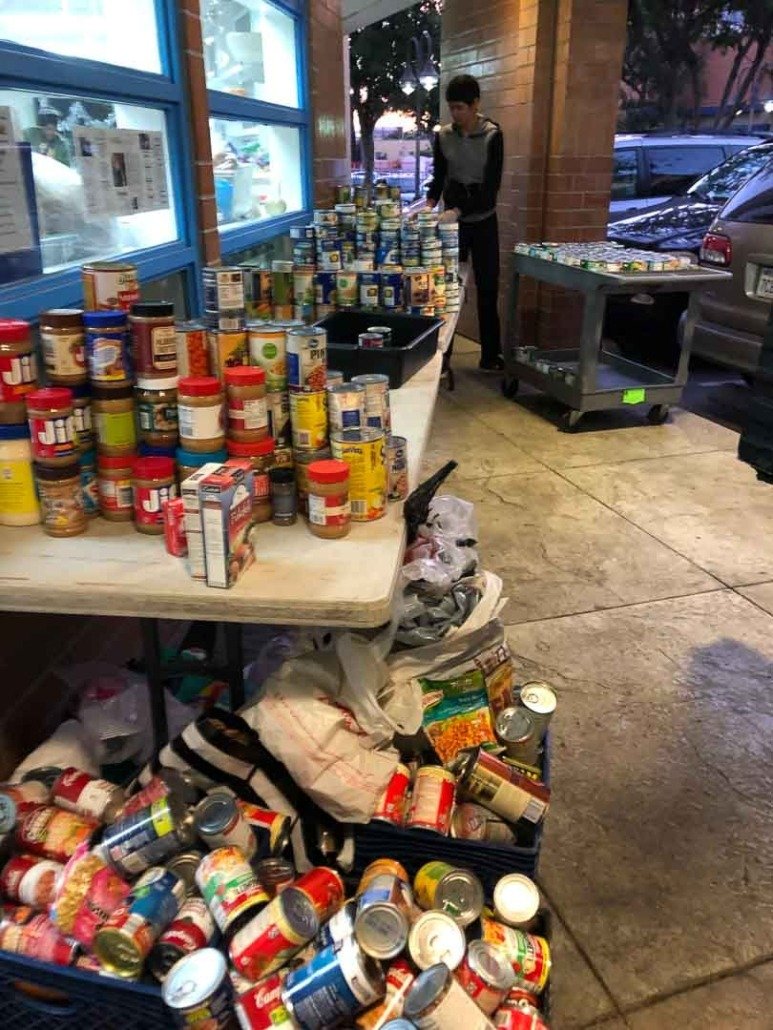 Table filled with cans and jars of food, with additional cans on the ground and in a black basket, outside of a building with brick and large blue windows.