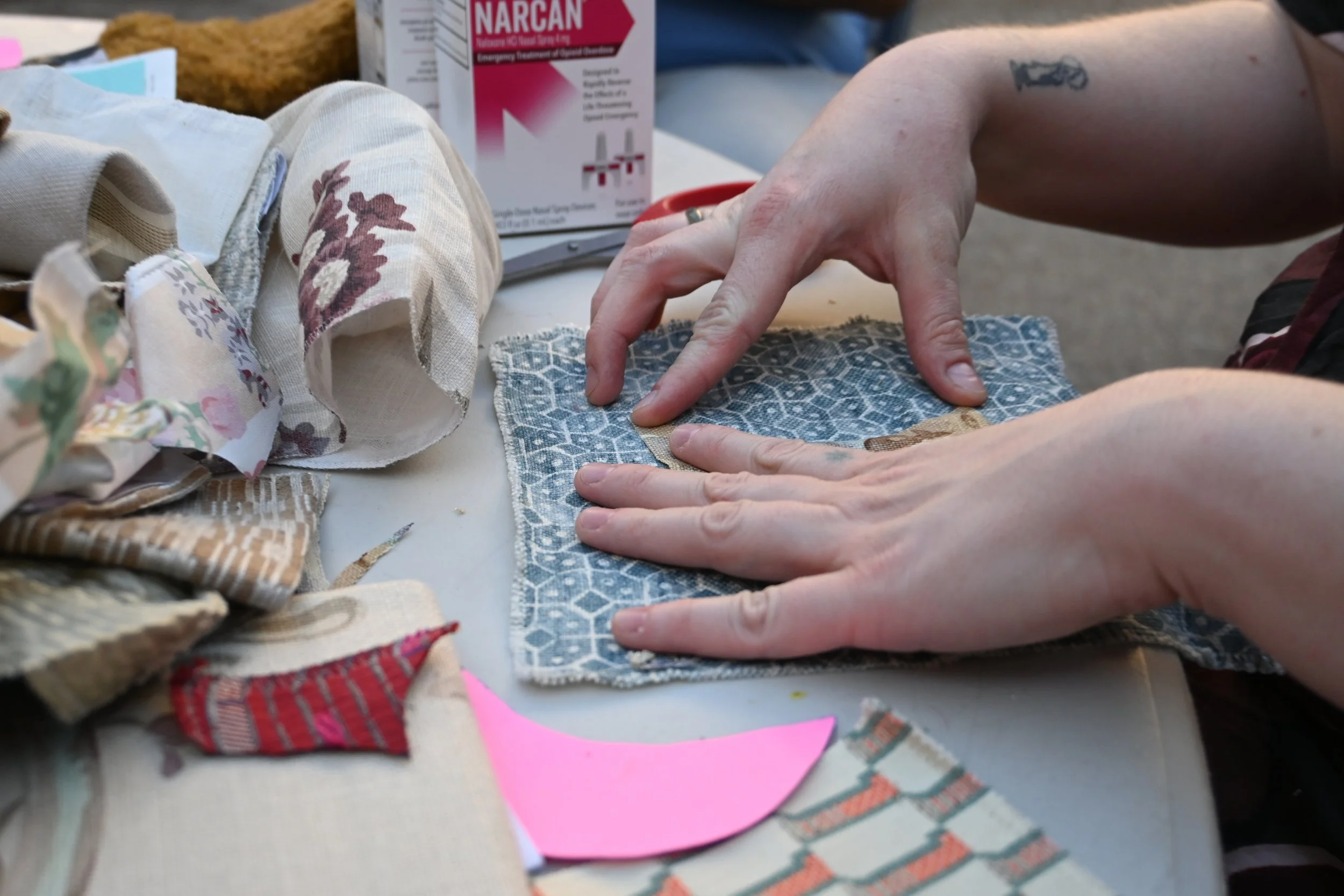 Hands folding a fabric square with a blue and white pattern at a sewing or crafting table, surrounded by various fabric scraps and materials.