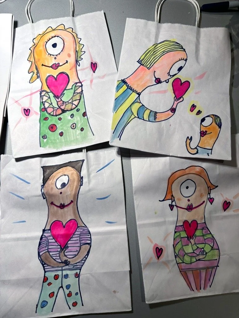 Four hand-drawn cartoon characters holding or surrounded by pink hearts, each with a large eye and colorful clothing, drawn on paper with clips at the top.