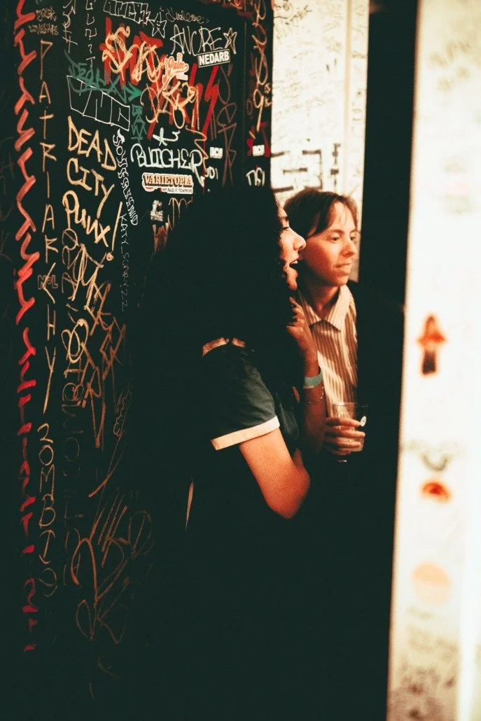 Two women conversing near a blackboard filled with colorful graffiti and signatures, one is smiling with curly hair and the other has straight hair, both appear to be in a casual setting.