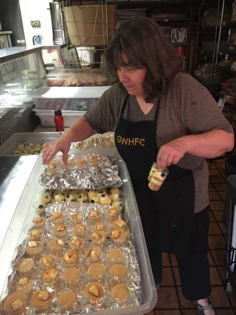 A woman working in a kitchen or food preparation area, placing small desserts into clear cups, with a tray of decorated desserts and foil-covered items on the counter.