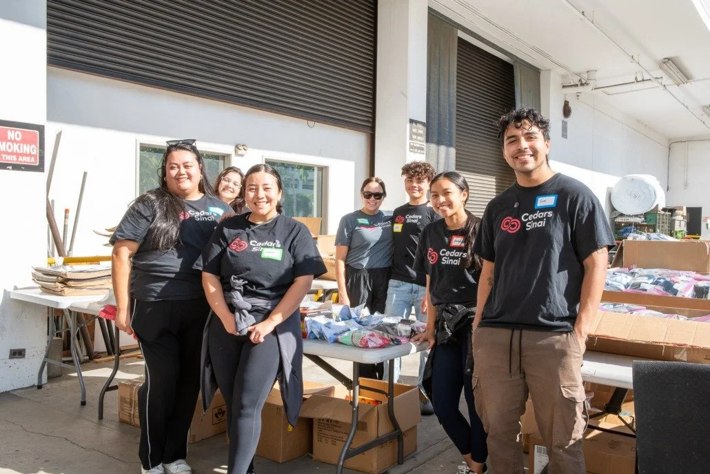 Group of seven people standing behind tables with clothes and supplies, smiling outdoors under a shelter.