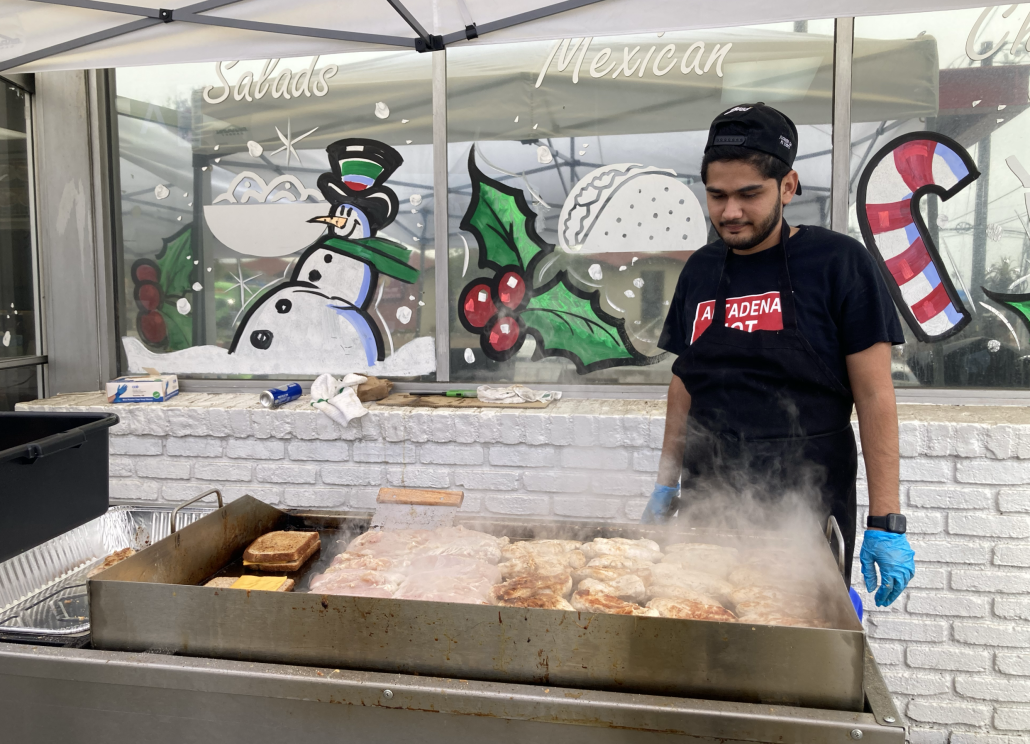 A young man cooking grilled chicken and other meats on a large outdoor grill outside a restaurant with Christmas-themed window art, including a snowman, holly, and candy cane.