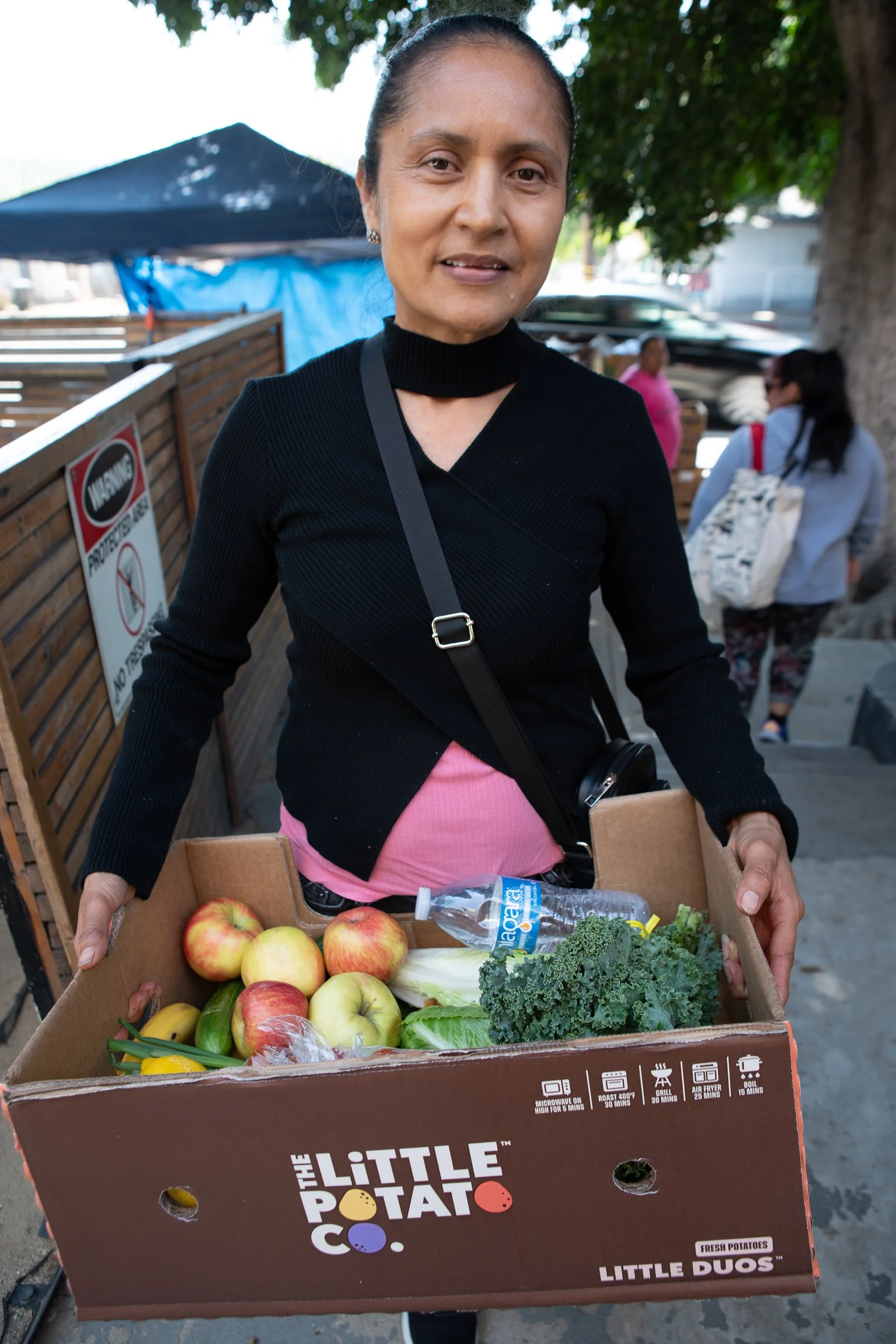 A woman holding a box of fresh produce, including apples, cucumbers, kale, and a water bottle, standing outdoors near a wooden fence and trees.