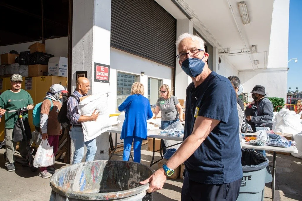 Man wearing glasses and a face mask placing trash into a large trash bin outdoors at a community event, with tables and people in line in the background.
