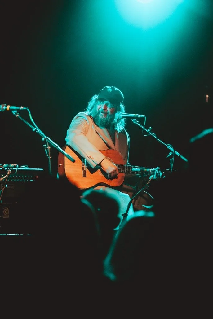 A musician with long hair, beard, glasses, and a cap, playing an acoustic guitar on stage with stage lighting, surrounded by microphones and musical equipment.