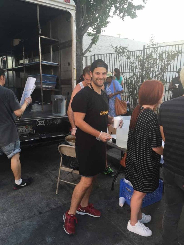 A smiling man wearing a black t-shirt, shorts, and red sneakers stands in line at an outdoor food truck event, holding a cup and wearing gloves. There are several people behind him, some waiting in line or preparing food. The scene takes place on a s