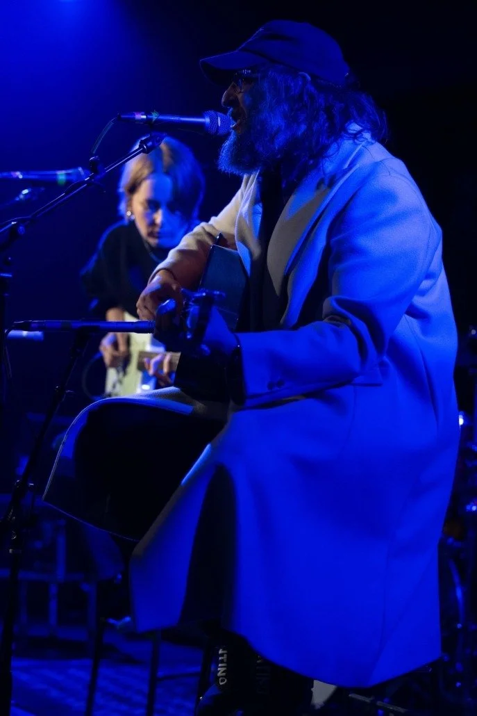 Musician wearing a hat and coat playing guitar and singing into a microphone during a live performance, with a woman in the background.