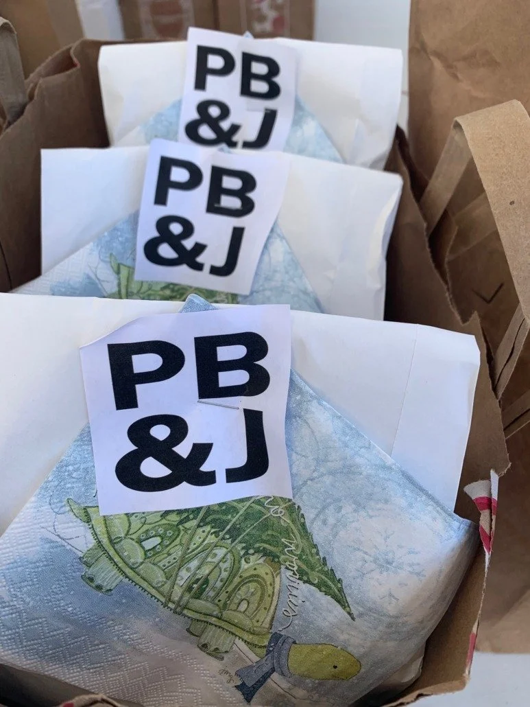 Three paper napkins each with a sticker labeled 'PB & J' placed on top inside a brown paper bag.