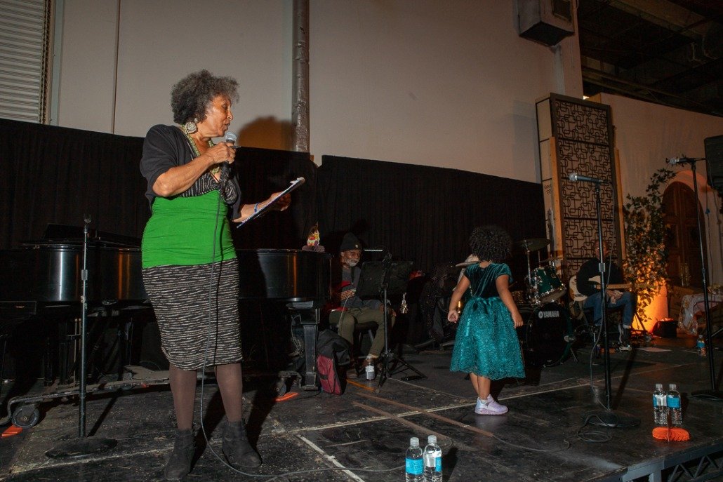 An older woman with gray curly hair, dressed in a black top with a green sash and a black-and-gray striped skirt, holding a microphone and script, standing on a stage. A three-piece band with percussion and keyboard instruments are behind her, and a 