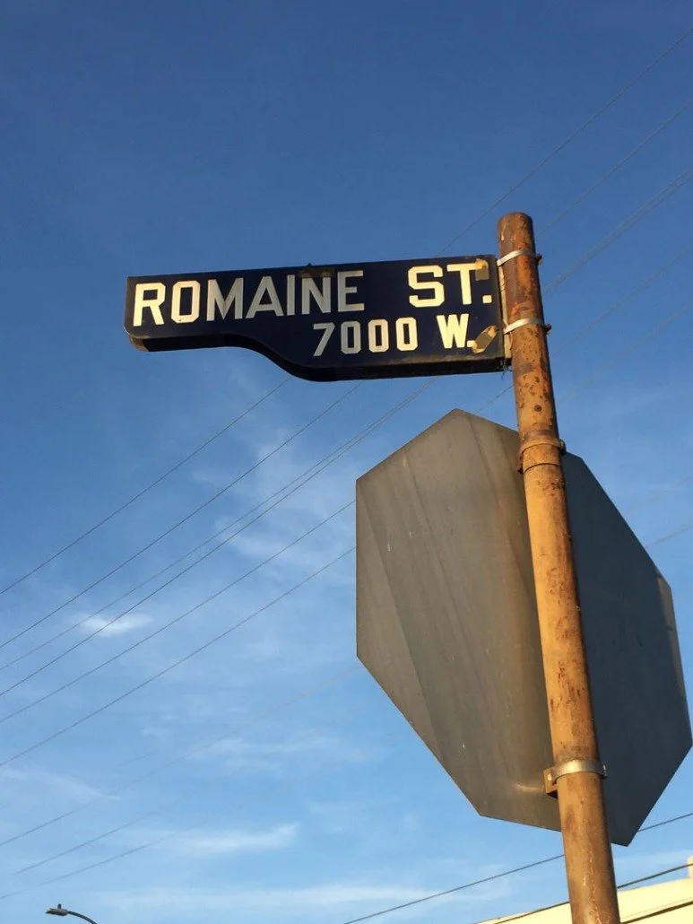Street sign reading ROMAINE STREET, with the address 7000 W, mounted on a rusted pole against a blue sky background.