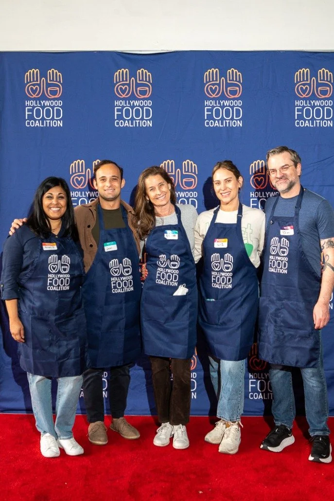 Group of five people standing in front of a blue backdrop with the 'Hollywood Food Coalition' logo, wearing dark blue aprons, posing and smiling.