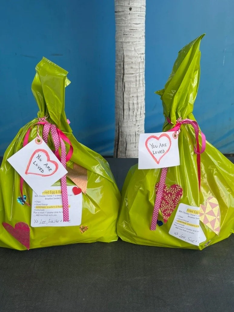 Two green gift bags decorated with pink ribbons and heart-shaped notes that read 'You Are Loved,' placed on a black surface with a blue wall and a tree trunk in the background.