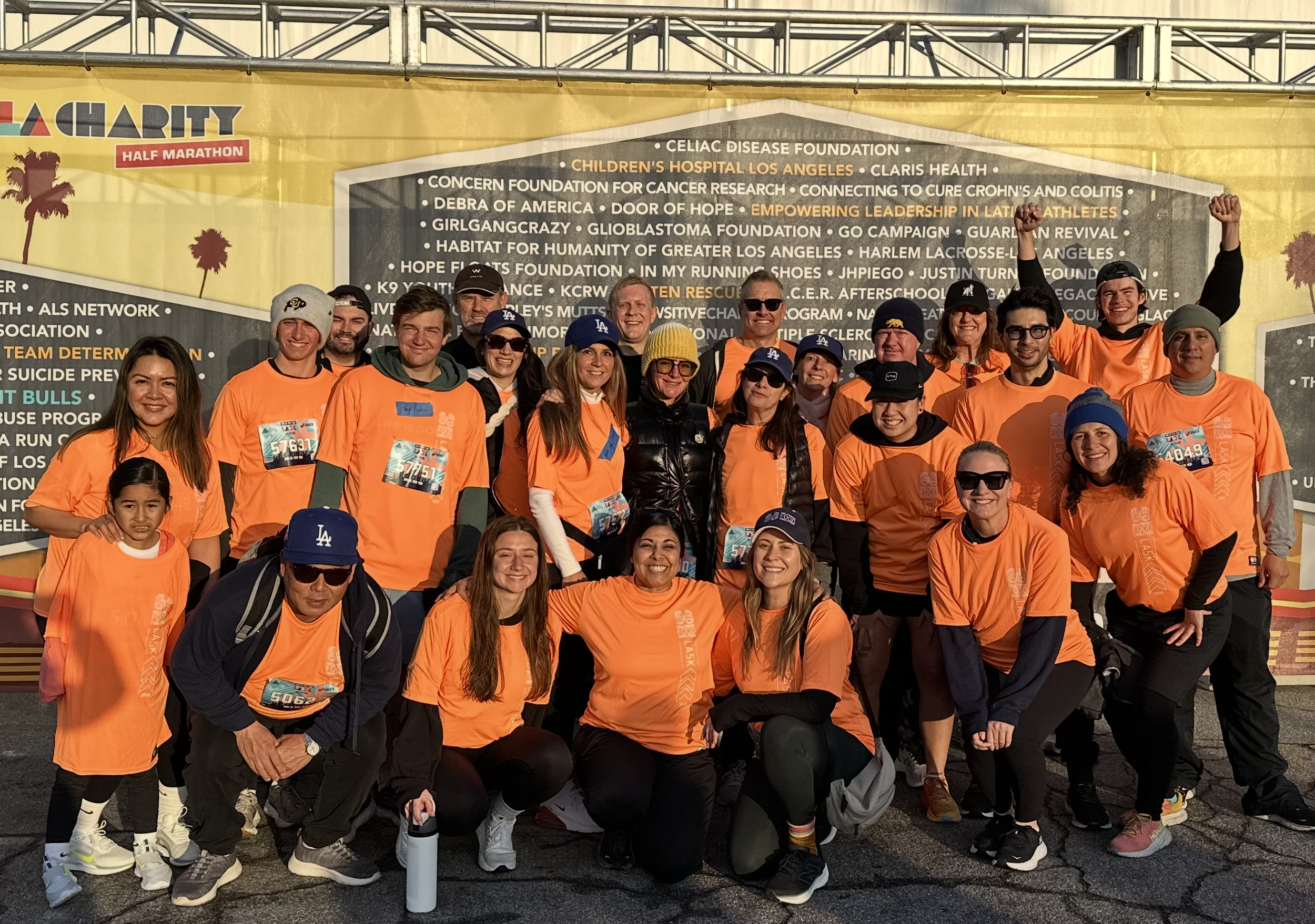 Group of people wearing bright orange shirts at charity event, some holding raised fists, standing in front of a large yellow banner with various foundations and organizations listed.