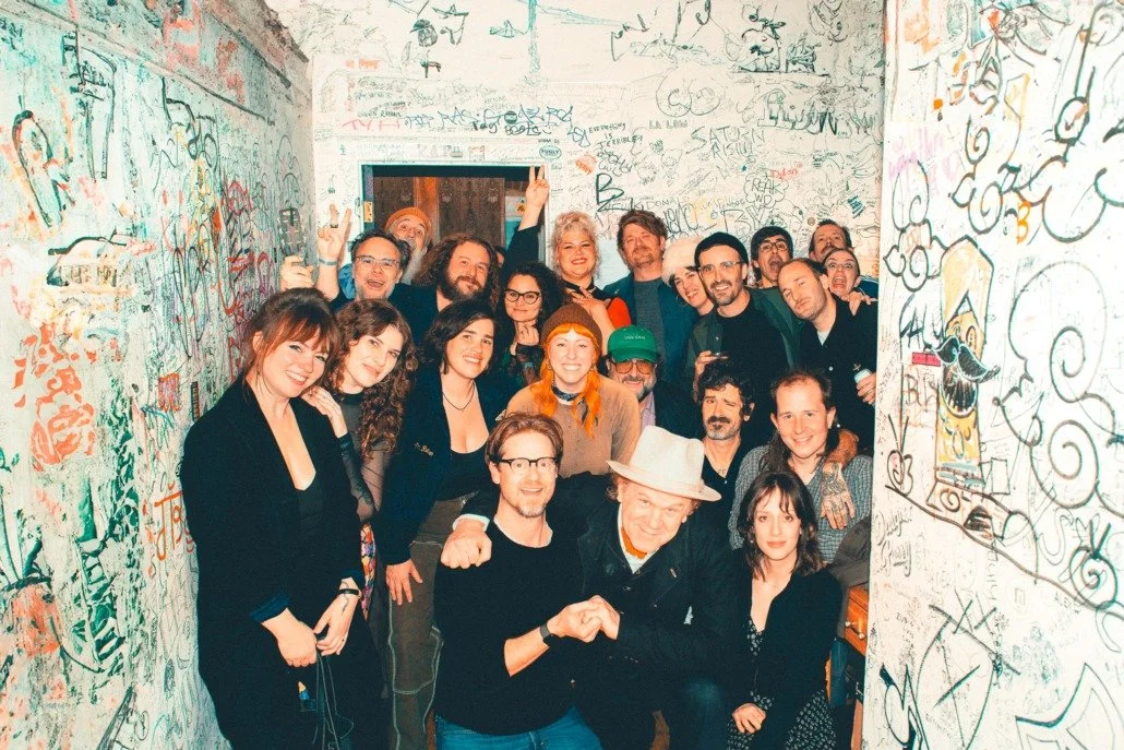 A group of people celebrating inside a graffiti-covered room, smiling, some making hand gestures, with a man in the center wearing a hat and a woman welcoming him with a handshake.
